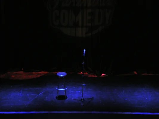 Scène vide pour un spectacle d'humour avec un tabouret et un micro sous un éclairage bleu.