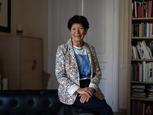 Helga Nowotny assise sur un canapé dans une pièce avec des bibliothèques.