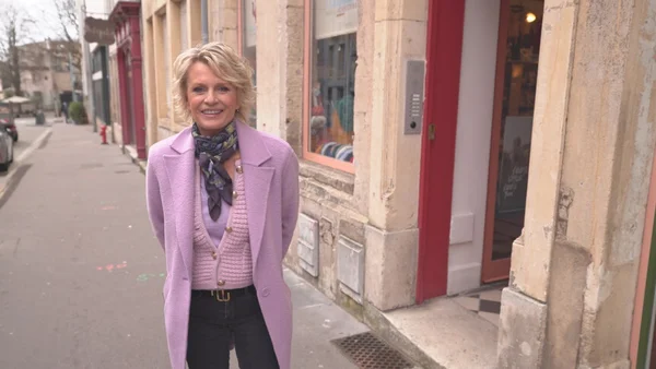 Sophie Davant devant une porte rouge, portant un manteau violet, un gilet rose et un foulard à motifs.