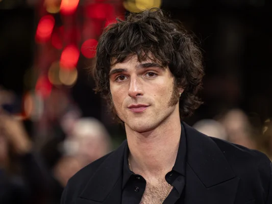 Jacob Elordi en costume trois-pièces marron et cravate noire.