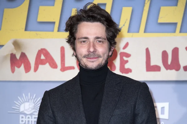 Guillaume Pley en col roulé et veste sombre, devant un fond coloré avec inscriptions.