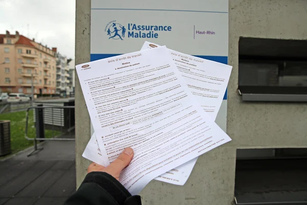 Une main tenant des arrêts maladie devant un panneau de l'Assurance Maladie en milieu urbain.