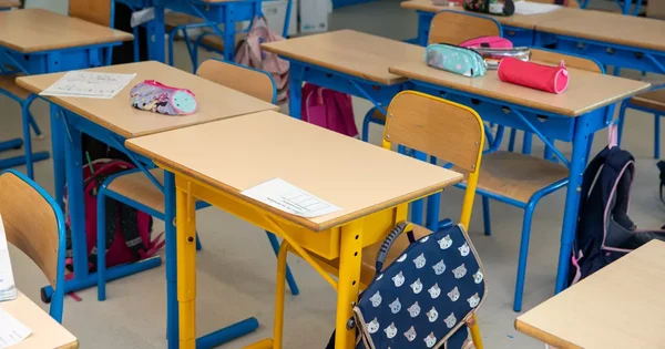 Une salle de classe vide avec des pupitres en bois, des chaises bleues et des sacs à dos rangés sous les tables.
