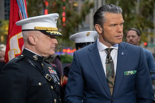 Pete Hegseth en tenue militaire aux côtés d'un civil en costume, lors d'un événement officiel en présence de drapeaux américains.
