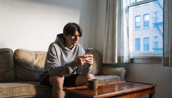 Jeune adulte assis sur un canapé dans un salon modeste, regardant son téléphone avec une expression hésitante, tenue de sport décontractée à côté