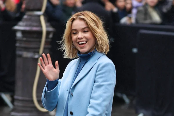 Milly Alcock saluant la caméra lors d'un événement public, vêtue d'un manteau bleu clair.