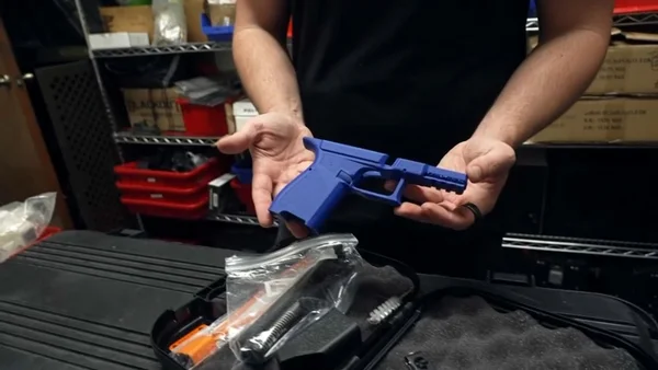 Une personne tenant un pistolet bleu imprimé en 3D au-dessus d'une mallette de pièces détachées dans un atelier.