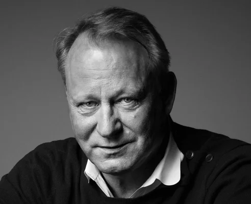 Portrait en noir et blanc de Stellan Skarsgård en pull sombre et chemise blanche.