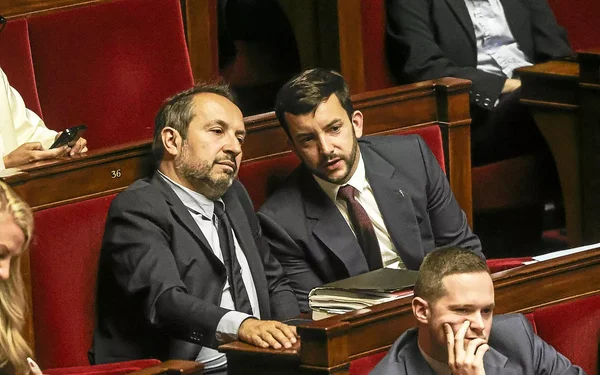 Deux hommes en costume dans l'hémicycle, en conversation dans un environnement parlementaire aux sièges rouges.