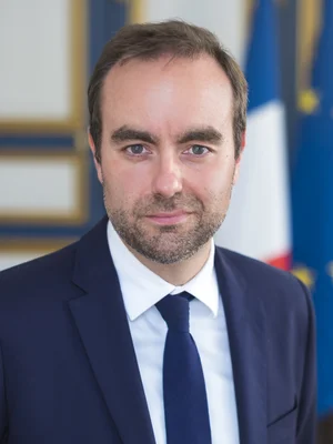 Sébastien Lecornu portant un costume bleu marine et une cravate foncée, posant devant un fond flou avec les drapeaux français et européens.