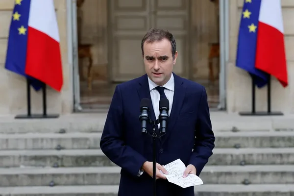 Sébastien Lecornu tenant des documents à l'extérieur d'un bâtiment officiel sous les drapeaux.