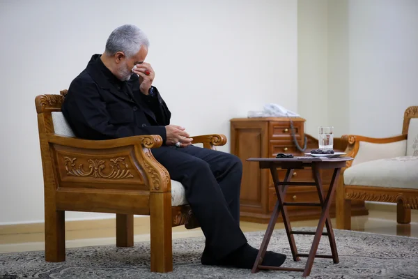 Qasem Soleimani assis dans un intérieur avec une table basse, essuyant son visage avec un mouchoir.