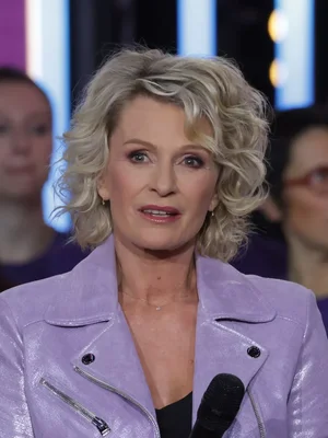 Sophie Davant en veste en cuir mauve tenant un micro, sur fond de figures floues.