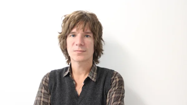 Portrait de Yann Destal en chemise à carreaux et gilet posant devant un fond blanc.