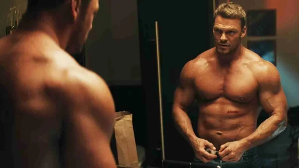 Alan Ritchson torse nu montrant sa musculature développée devant un miroir.