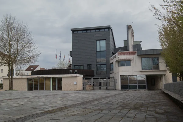 Un bâtiment moderne avec une place pavée et des drapeaux, sous un ciel couvert.