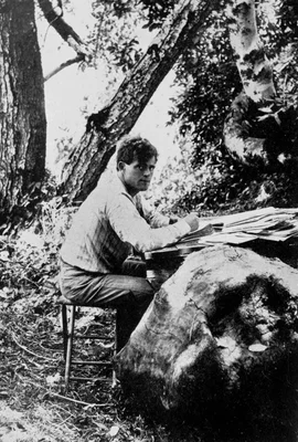 Jack London écrivant en plein air en 1905, assis devant une table de fortune au milieu des arbres.