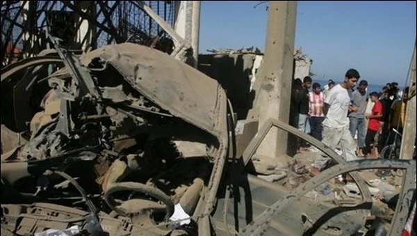 Véhicule détruit et bâtiment endommagés suite à une attaque, avec des secours sur place.