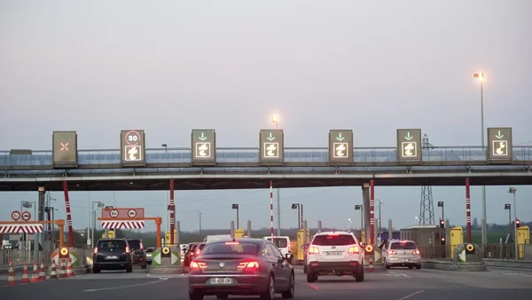 Véhicules attendant aux cabines de péage d'une gare autoroutière avec panneaux électroniques et barrières.