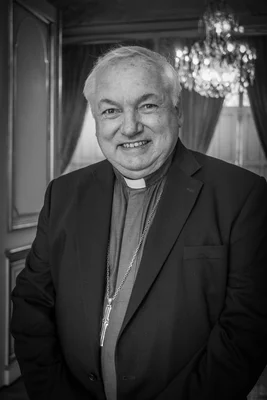 Mgr Jean-Marc Aveline portant une chemise avec col romain et une chaîne avec croix, photographié dans un intérieur élégant.