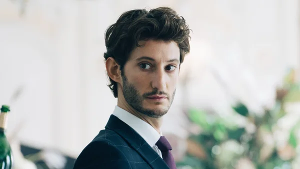 Pierre Niney en costume à carreaux et cravate violette, regardant sur le côté.