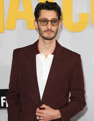 Pierre Niney en blazer bordeaux et lunettes, tenant le revers de sa veste.