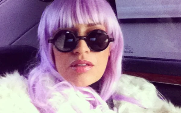 Laam aux cheveux violets portant des lunettes de soleil et un manteau de fourrure blanche, assise dans une voiture.