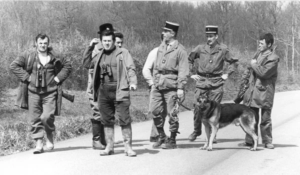 Six hommes et un chien berger allemand, dont trois militaires, sur une route rurale lors d'une patrouille de recherche.