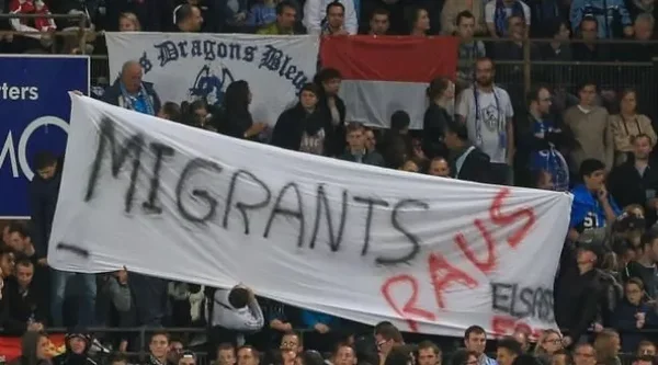 Supporters au stade exhibant une banderole 'MIGRANTS RAUS' avec mention 'ELSASS'.