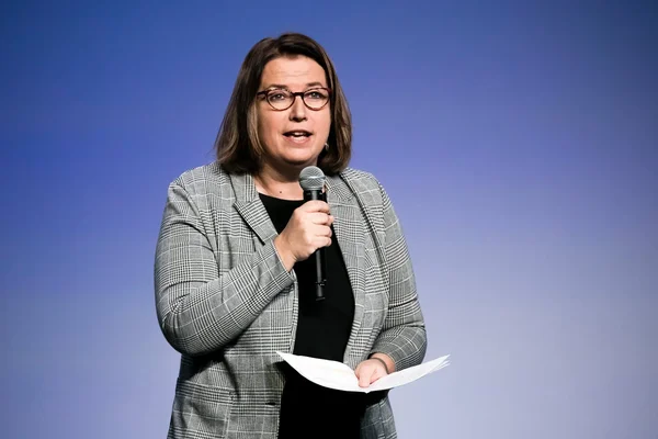 Claire Hédon, Défenseure des droits, s'exprimant devant un micro avec un blazer gris à carreaux sur fond bleu.