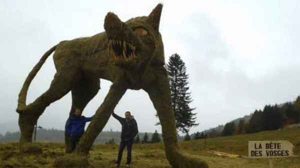 Créature en mousse figurant la Bête des Vosges, exposée dans un champ avec deux personnes posant devant elle.