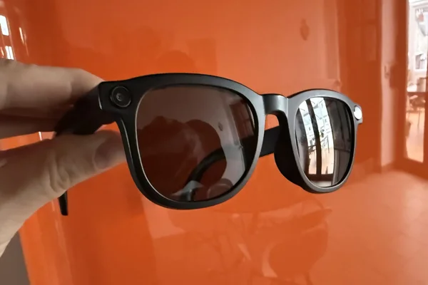Une main tient les lunettes connectées Blacksheep à monture noire, posées sur un fond orange.