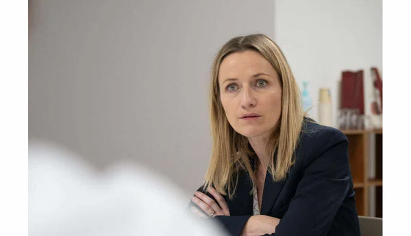 Femme blonde en veste sombre, assise bras croisés dans un intérieur avec des étagères en arrière-plan.