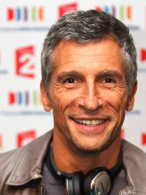 Nagui souriant en veste en cuir, portant des écouteurs, devant un fond coloré aux logos de la chaîne.
