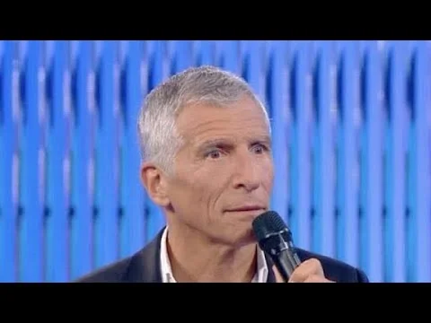 Nagui micro en main sur un plateau avec un fond bleu rayé.