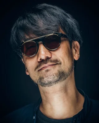 Hideo Kojima posant avec des lunettes de soleil et une tenue sombre.