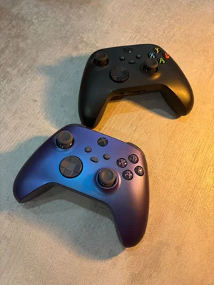 Deux manettes Xbox, l'une noire et l'autre bleu-violet, posées sur une surface claire.