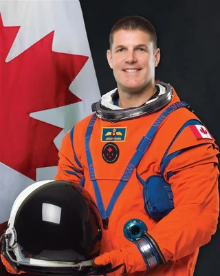 L'astronaute Jeremy Hansen en combinaison orange tenant un casque devant le drapeau canadien.