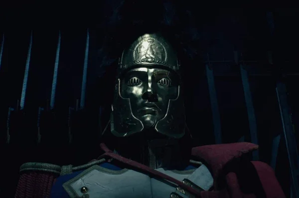 Un personnage en armure détaillée portant un casque à emblème et une cape rouge dans un environnement sombre.