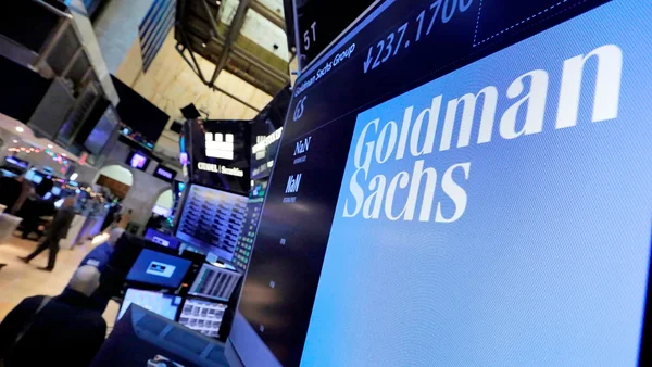 Écran numérique affichant le nom Goldman Sachs sur fond bleu, dans un contexte de salle de marchés financiers.