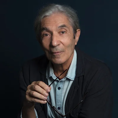 Boualem Sansal tenant ses lunettes devant un fond sombre.