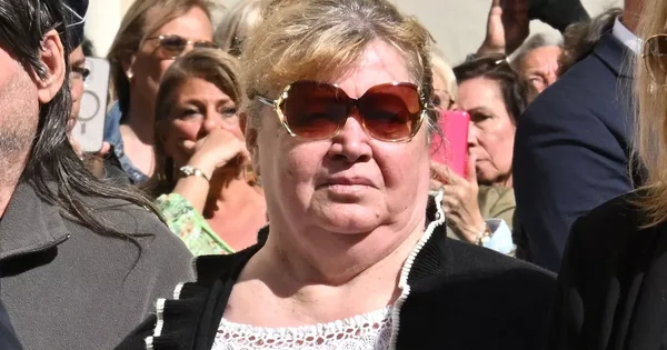 Une photo montre une femme aux cheveux blondes portant de grands lunettes de soleil bruns et une veste noire sur un haut en dentelle blanche, debout parmi une foule de personnes qui semblent être à l'extérieur en plein jour. Certains individus à l'arrière-plan tiennent des téléphones en l'air, suggérant un événement public ou un rassemblement.