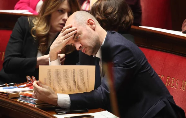 Un homme en costume assis à un bureau dans un environnement législatif, tenant un document.