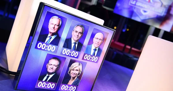 Un écran de télévision affichant cinq candidats en costume avec des minuteurs dans un studio de télévision