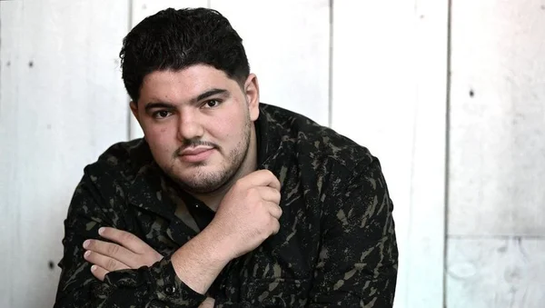 Mehdi Kessaci posant devant un mur en bois blanc, vêtu d'une chemise camouflage.