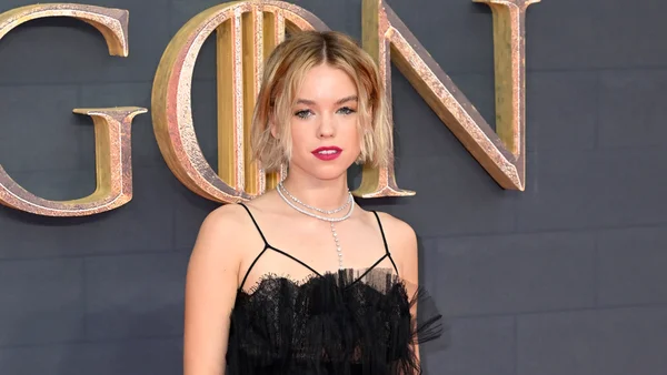 Milly Alcock en robe noire en dentelle et colliers de perles devant un mur aux lettres dorées.