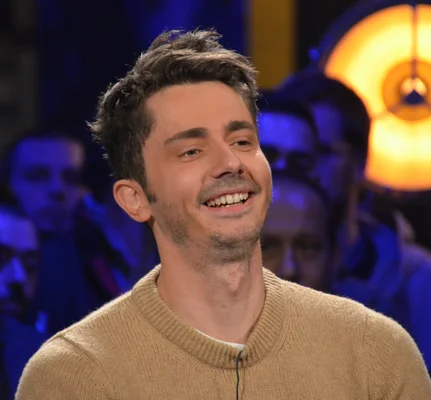 Guillaume Pley souriant en pull beige tricoté, devant un public flou et un luminaire circulaire.