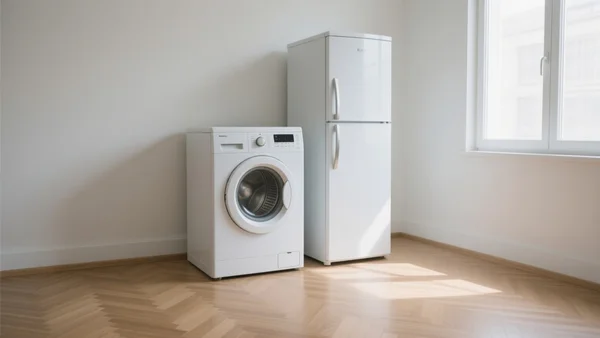 Petit réfrigérateur blanc et lave-linge frontal posés côte à côte dans un studio vide avec parquet clair, lumière naturelle provenant d'une fenêtre