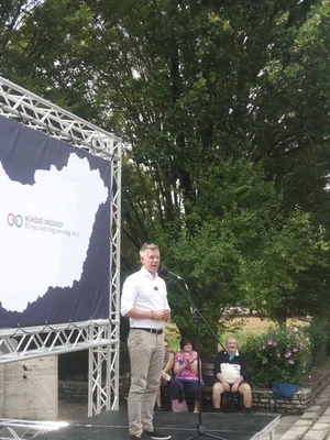 Péter Magyar tenant un discours sur scène à Battonya, devant une bannière électorale et une nature estivale.