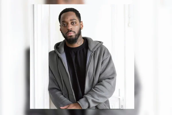 Flavel, porteur d'un sweat gris, posant les mains jointes devant un fond clair pour témoigner.